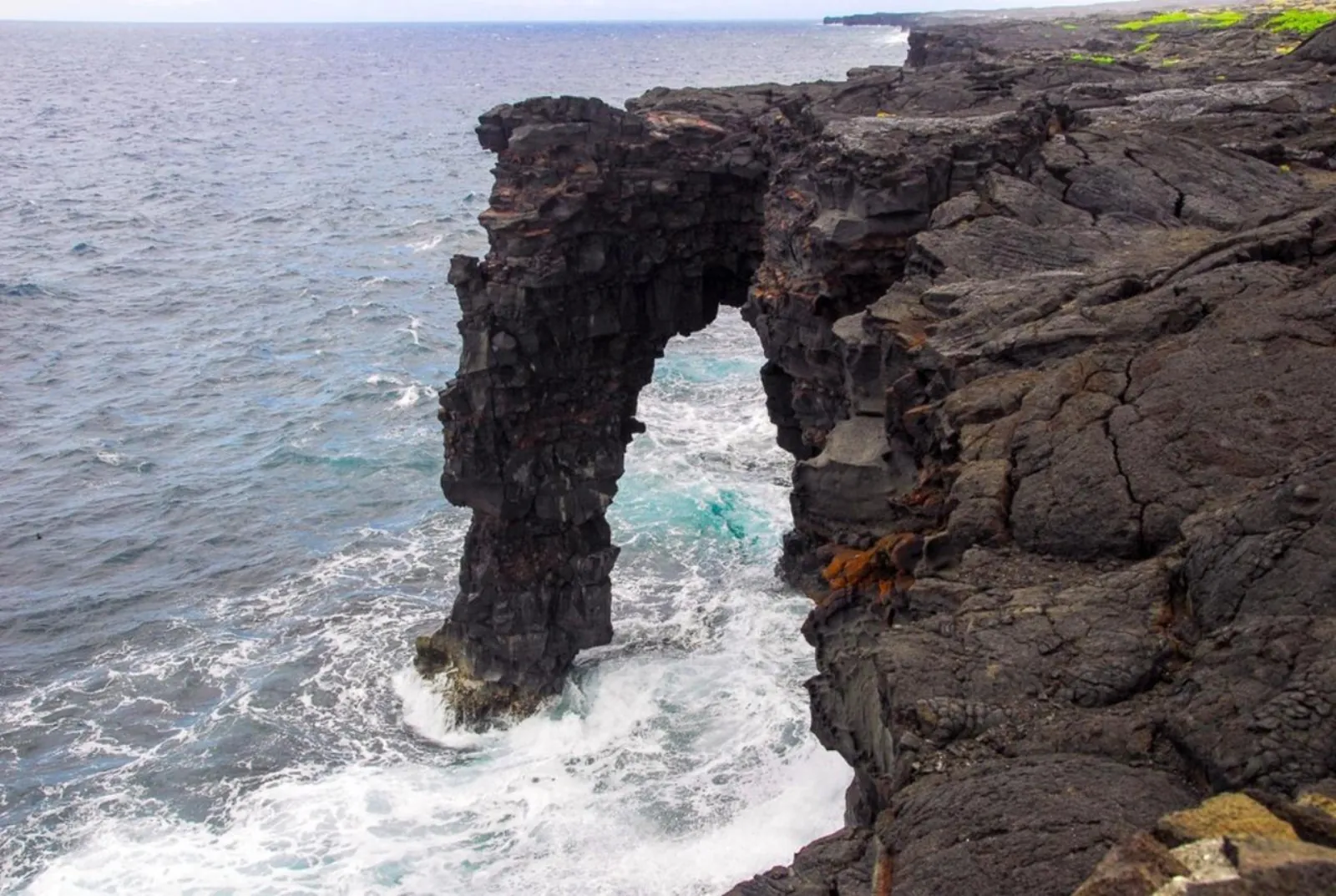 A tall black lava sea arch with waves breaking through it below a rocky cliff