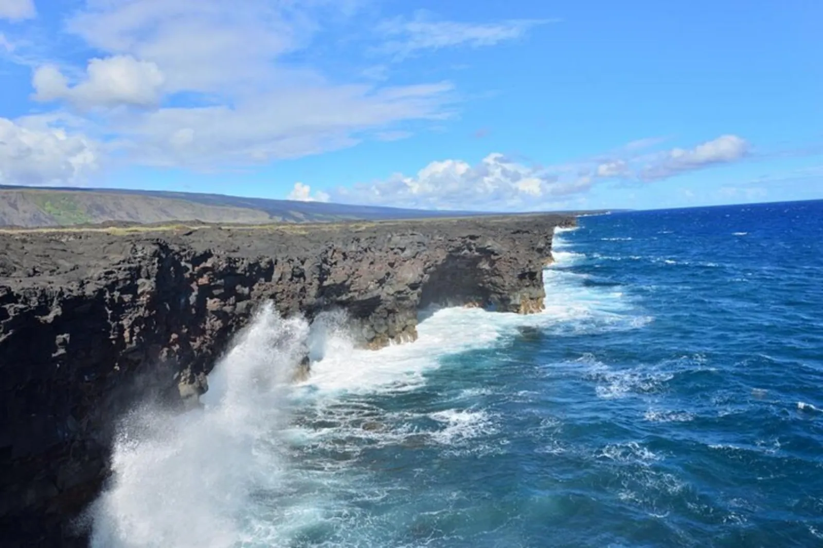 Waves crash against dark lava cliffs along a coastline under a blue sky