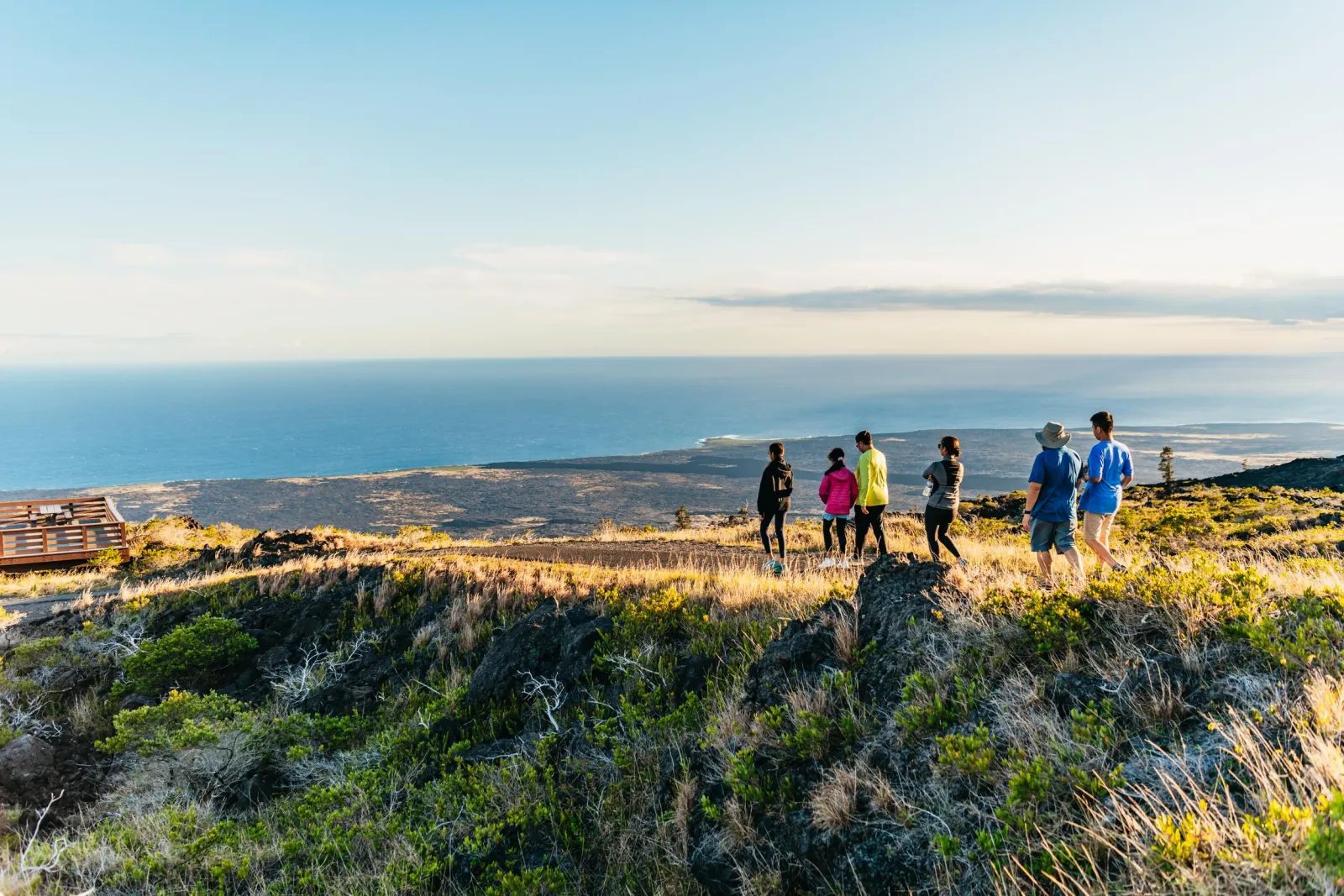 Small group walking a grassy lava slope above a wide coastal plain and ocean