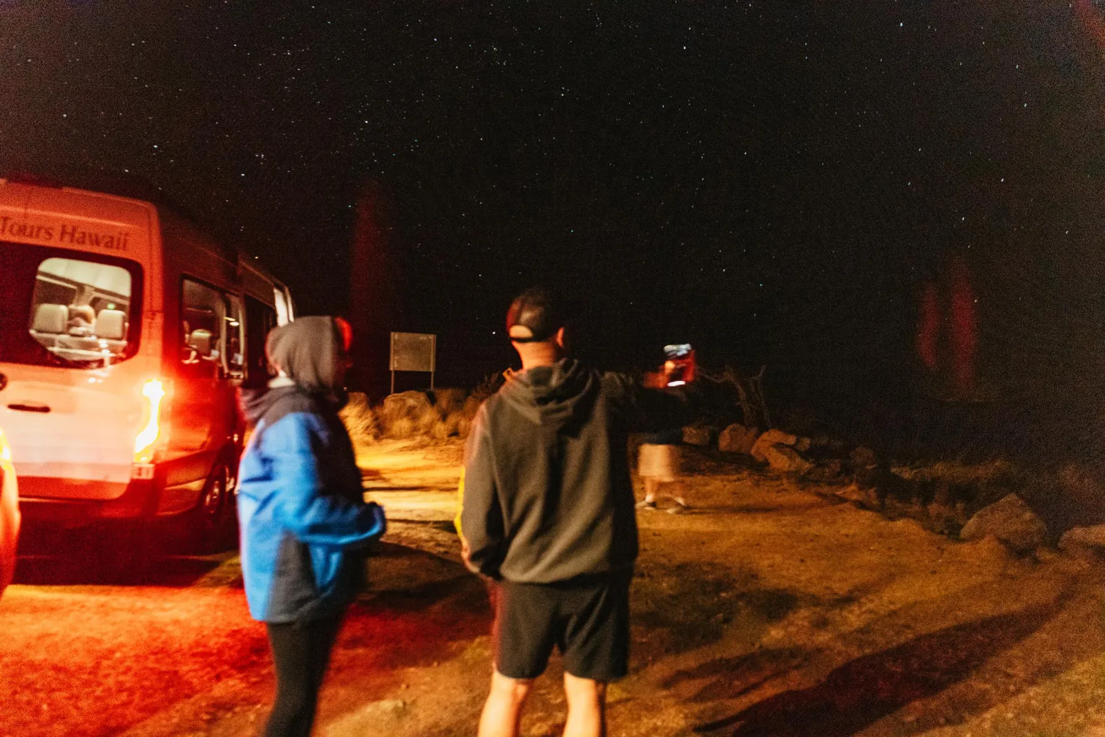 Guests in hoodies photographing a starry sky beside a tour van at night