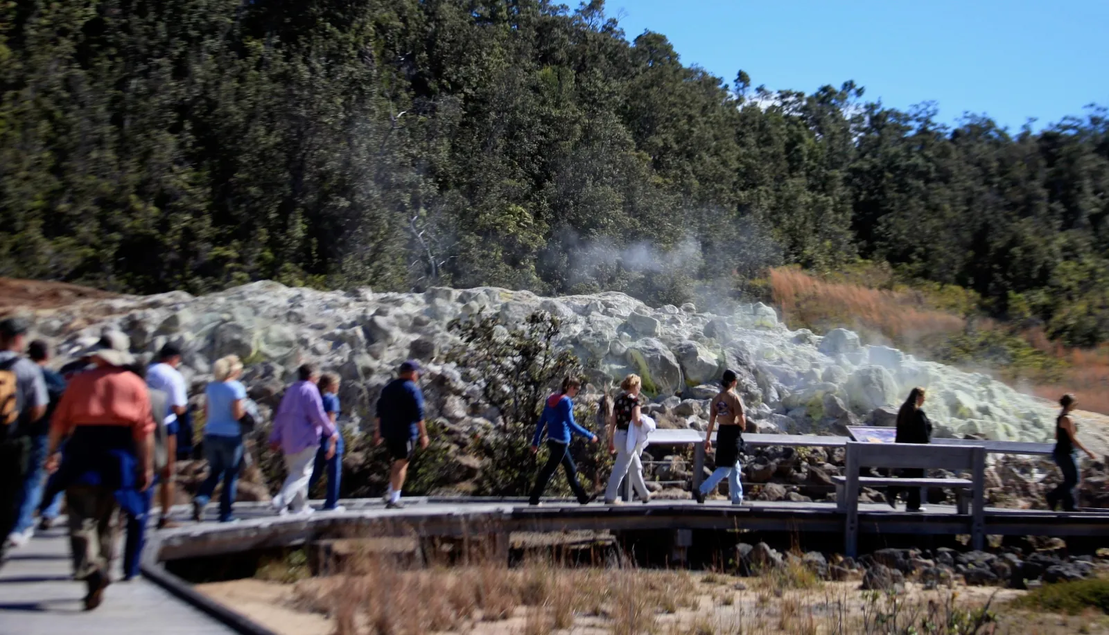 Line of visitors on a wooden boardwalk past steaming yellow-white sulfur rocks