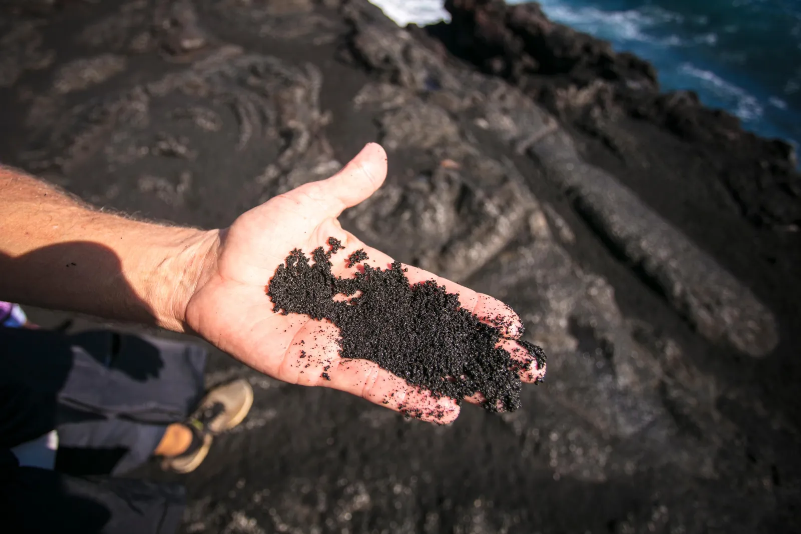 Hand holding coarse black sand on a lava shoreline above the blue ocean