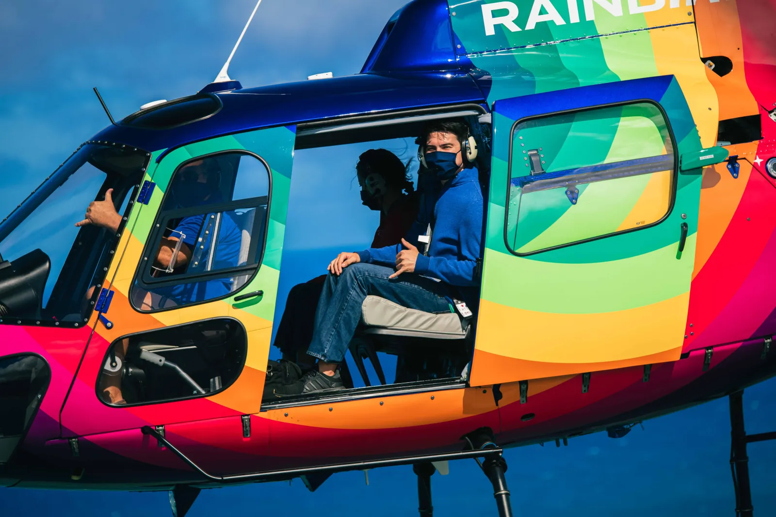 Passengers with headsets seated in a rainbow-painted helicopter flying with its door off