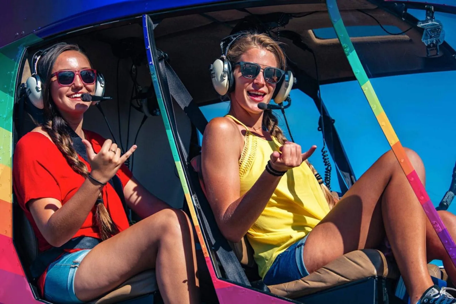 Two smiling passengers with headsets and sunglasses in an open-door rainbow helicopter