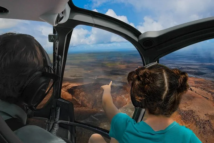 Passenger in a helicopter cockpit pointing down at a reddish volcanic crater landscape