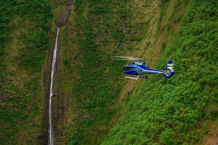 Blue helicopter flying beside a tall, thin waterfall on a steep green valley wall