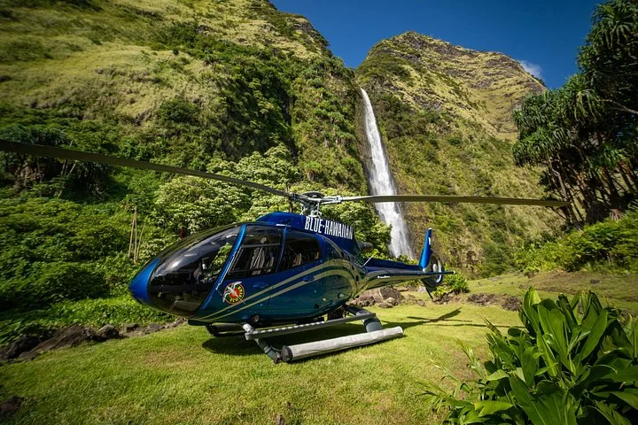 Blue Hawaiian helicopter parked on a grassy pad below a waterfall and green cliffs
