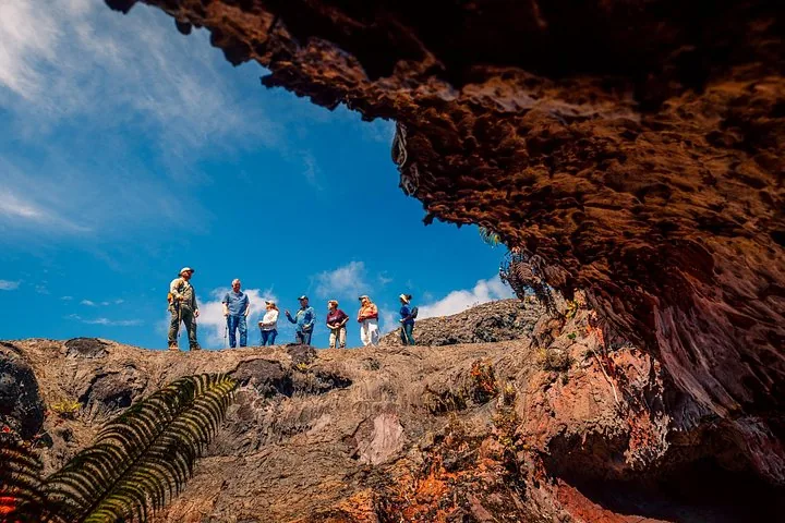 Hikers standing on a lava ridge under blue sky, framed by a rocky overhang and fern