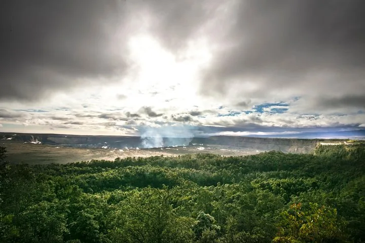 Steam plume rising from a wide summit caldera beyond green forest under storm clouds