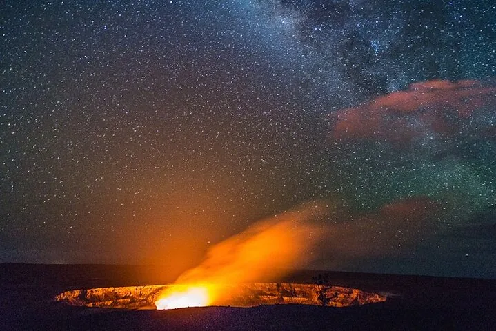 Orange glow and plume rising from a crater rim beneath a starry Milky Way