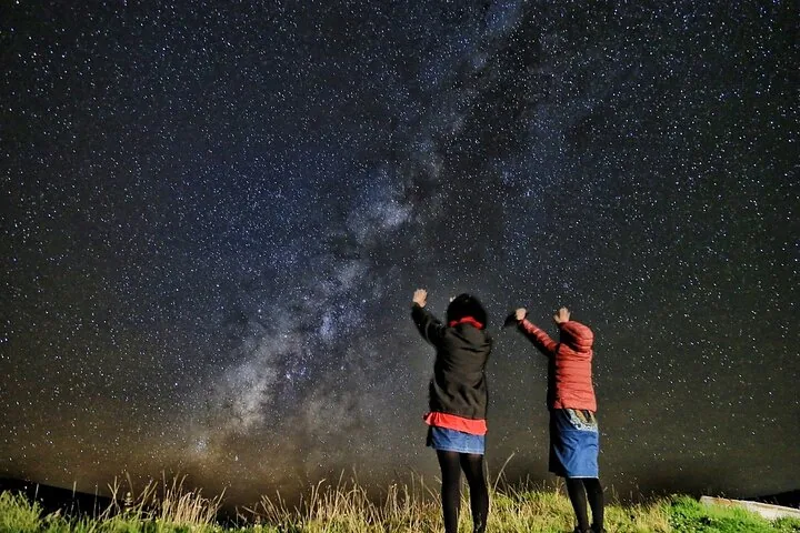 Two guests raising their arms beneath the Milky Way on a grassy hillside at night