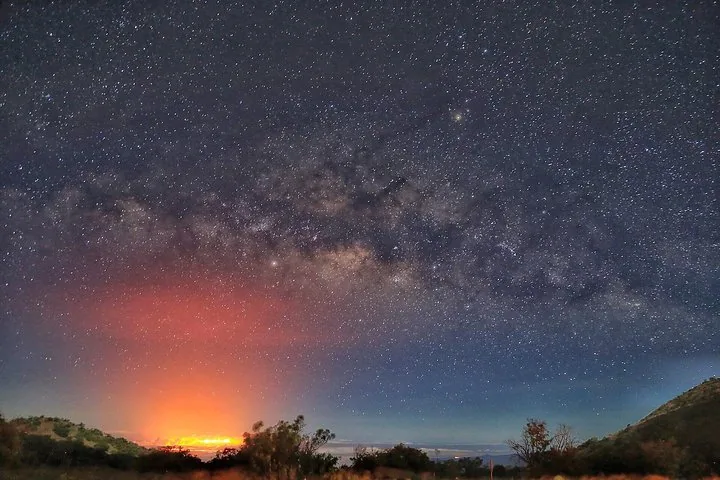 Milky Way arching over a dark ridge with an orange glow on the horizon