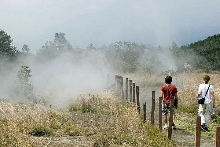 Two visitors walking a fenced path through steam rising from grassland vents