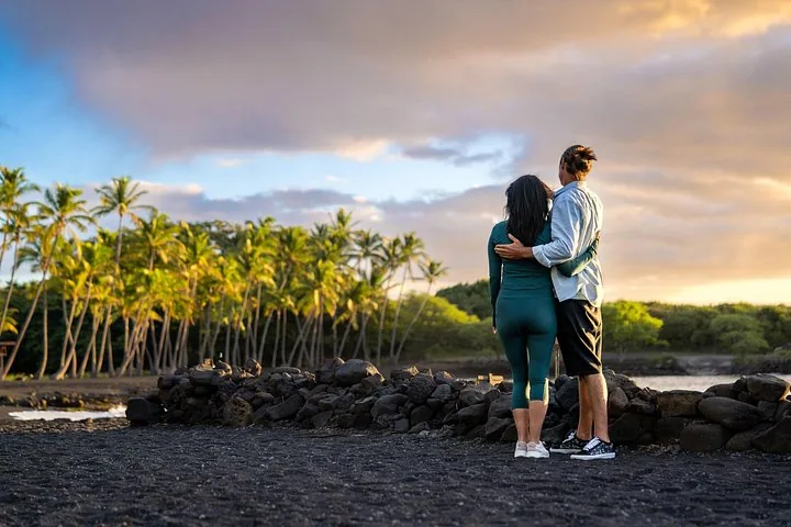 Couple embracing on a black sand shore at sunset, coconut palms behind a stone wall